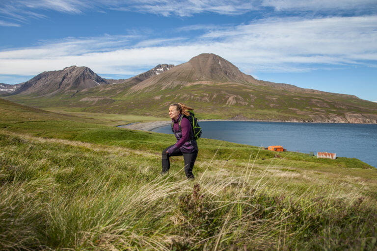 Kuvassa nainen patikoi Islannissa kesäaikaan päällään violetti paita, mustat housut ja selässä retkeilyreppu. Taustalla näkyy vuoria ja merenlahti.